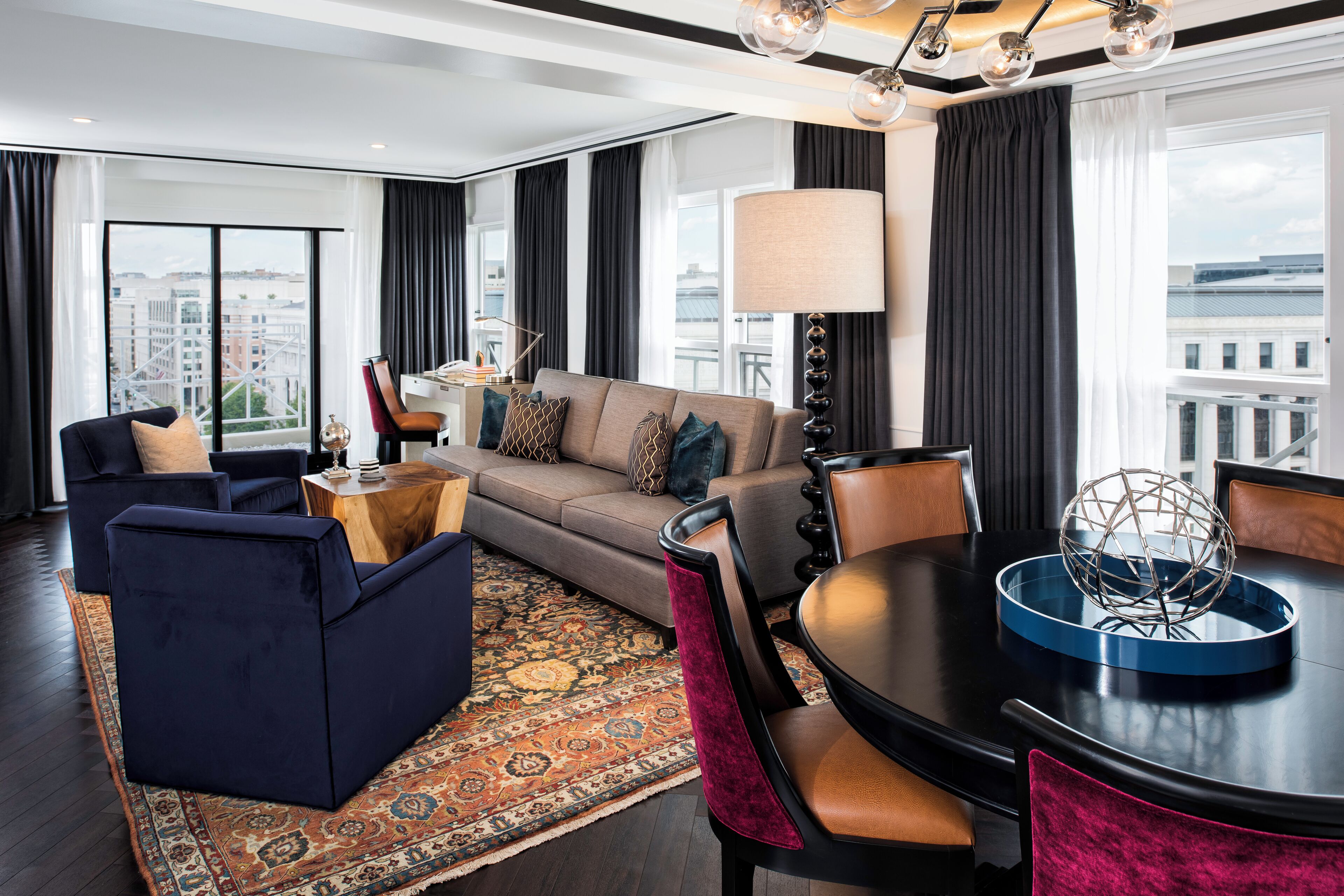 A modern hotel room with a sofa, chairs, a dining table, and a large window offering a city view.