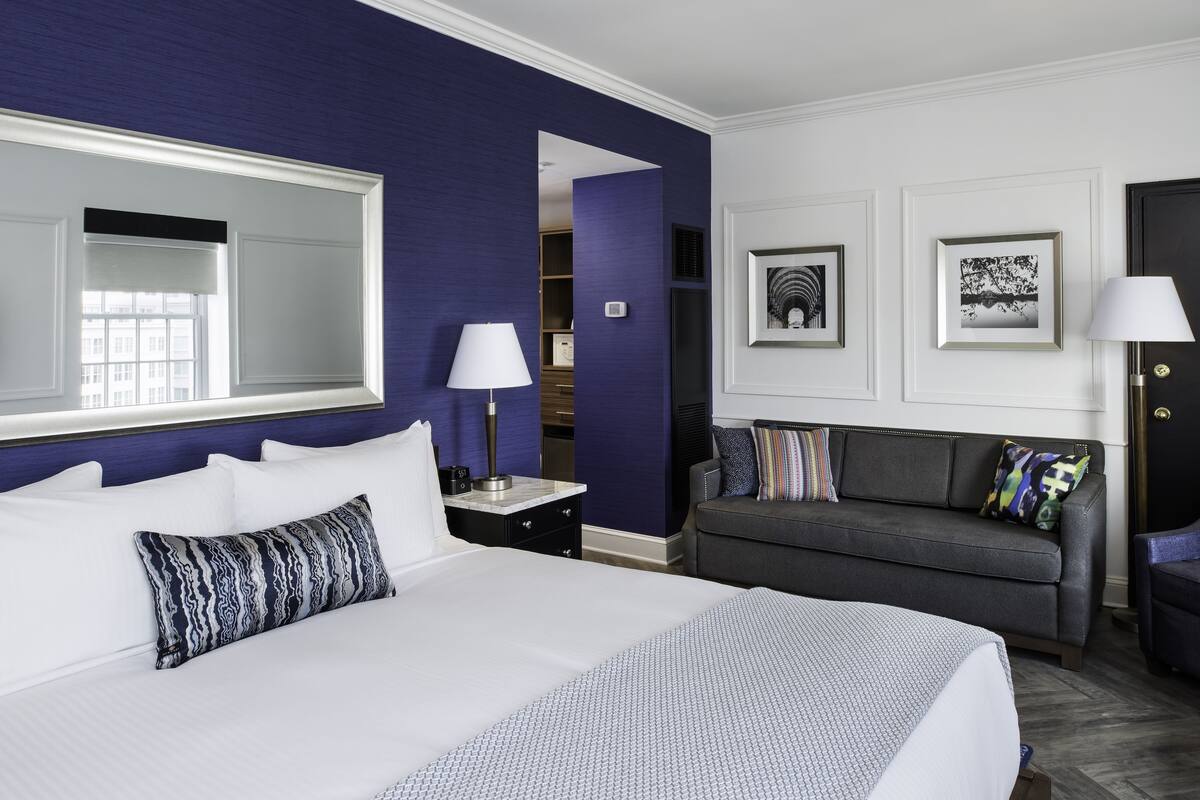 A hotel room with a large bed, a grey sofa, a mirror, and two framed pictures on the wall.