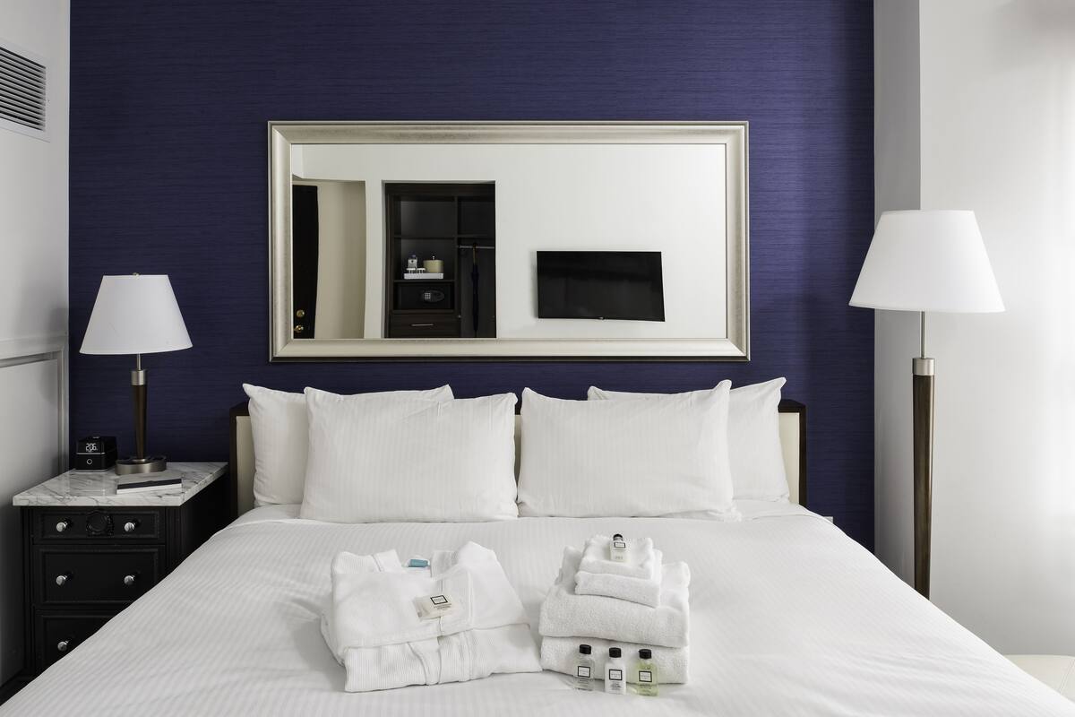 A neatly made bed with white linens, a large mirror above it, and a bedside table with a lamp and a clock.