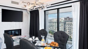 Una habitación de hotel moderna con una mesa de comedor de vidrio, sillas tapizadas en negro, un televisor de pantalla plana, una chimenea y vistas a la ciudad.