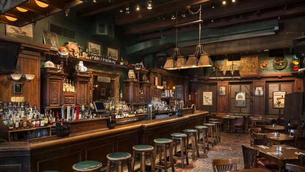 Un área de bar con mostradores de madera, taburetes y diferentes botellas y vasos.