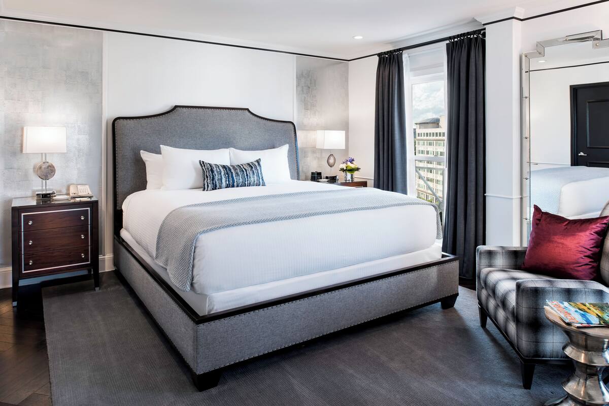 A modern hotel room with a large bed, a nightstand, a chair, and a view of the city.