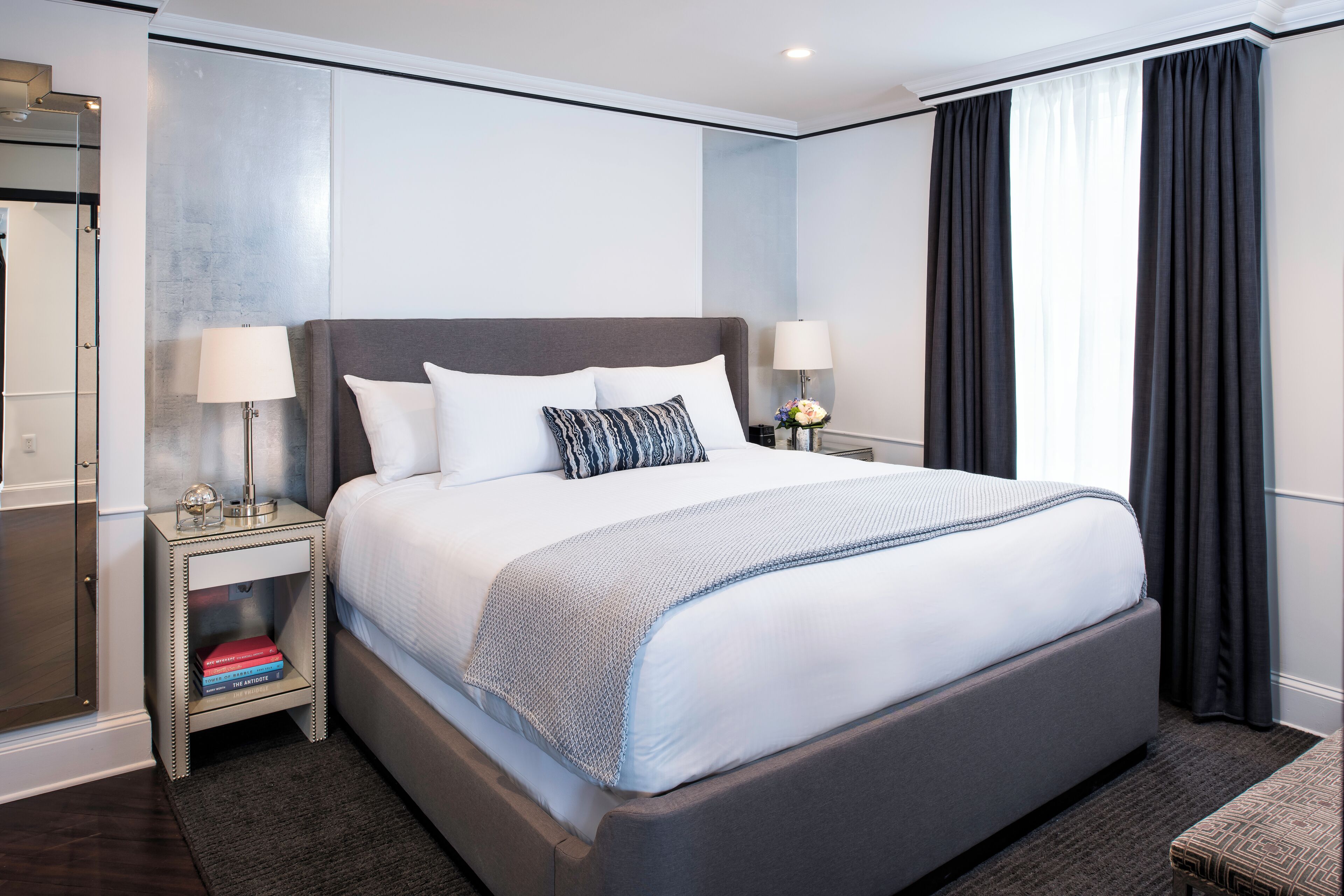 Une chambre d’hôtel moderne avec un grand lit, des lampes de chevet, une table de chevet avec des livres et une vue sur l’extérieur par une fenêtre munie de rideaux.