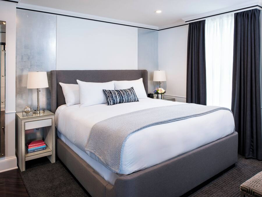 A modern hotel room with a large bed, bedside lamps, a nightstand with books, and a view of the outside through a window with curtains.