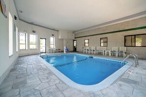 Una piscina interior con suelo de piedra, paredes blancas y una franja verde. Se observan sillas y una puerta con un cartel.
