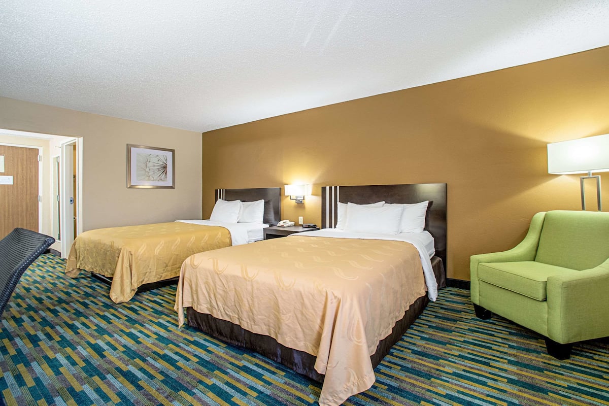A hotel room with two beds, a green armchair, a patterned carpet, and a framed picture on the wall.