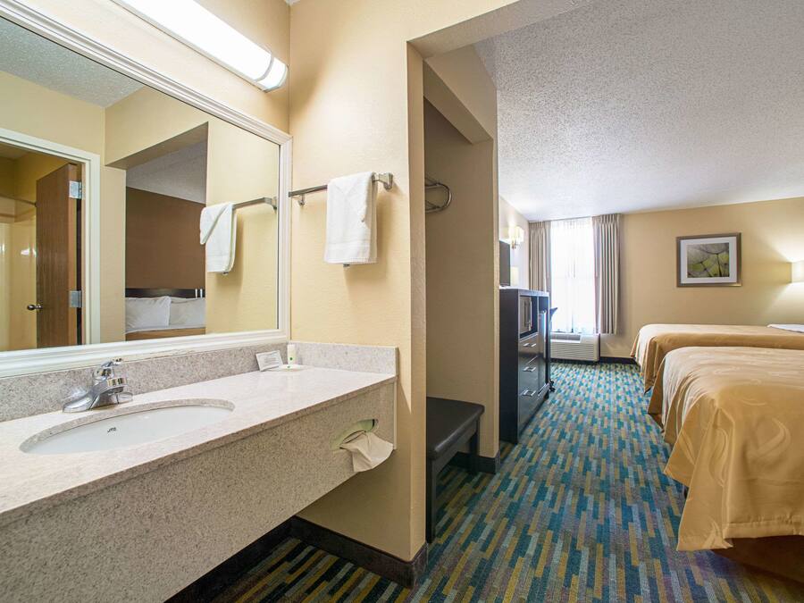 A hotel room with a large bathroom mirror, a sink, a bench, and two beds with beige bedding.