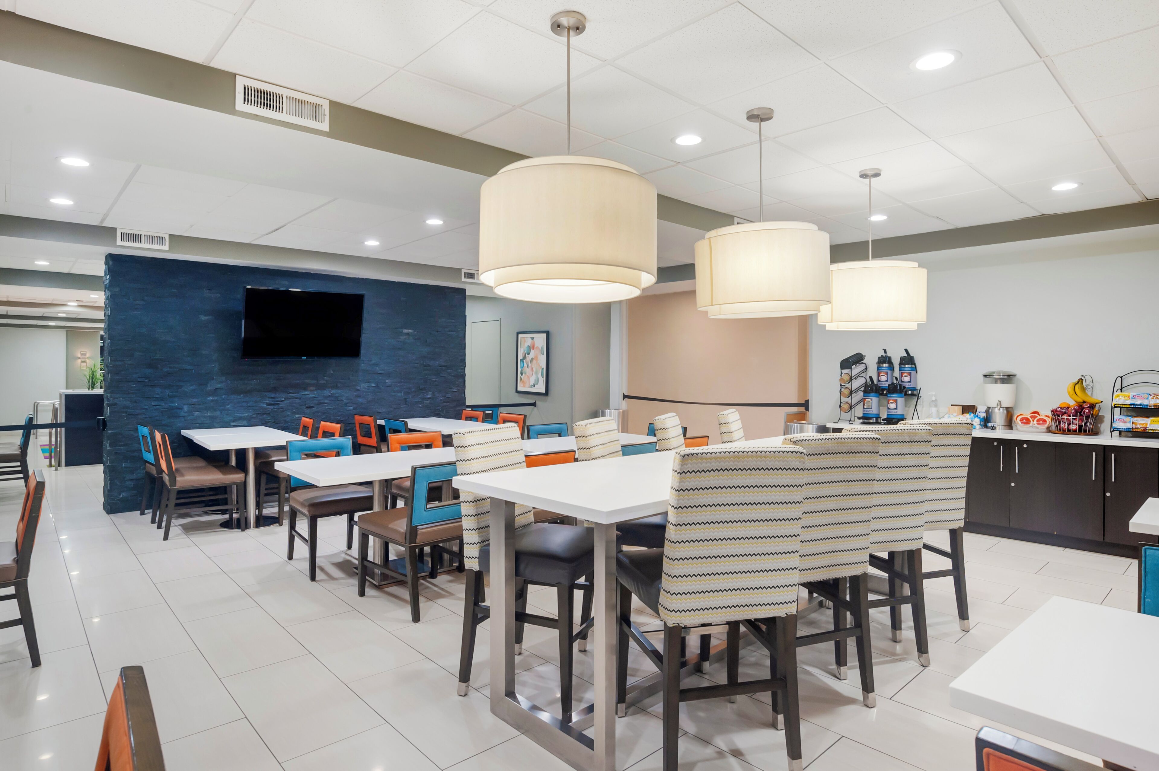 Zona pranzo moderna con tavoli e sedie, una TV a schermo piatto montata su una parete blu, illuminazione a sospensione e un bancone cucina con diversi oggetti.