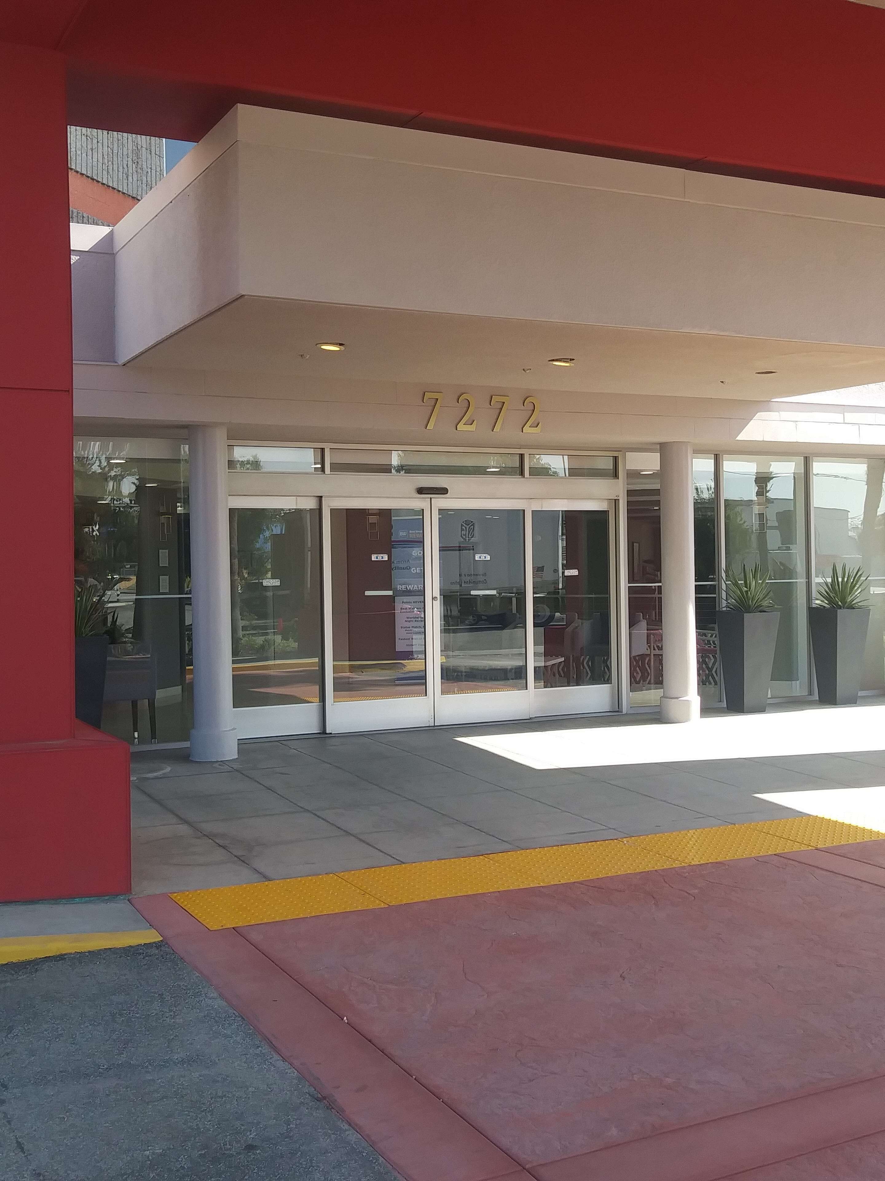 A modern building entrance with a glass door, a number '7272' above, and a clear view of the interior through the glass.