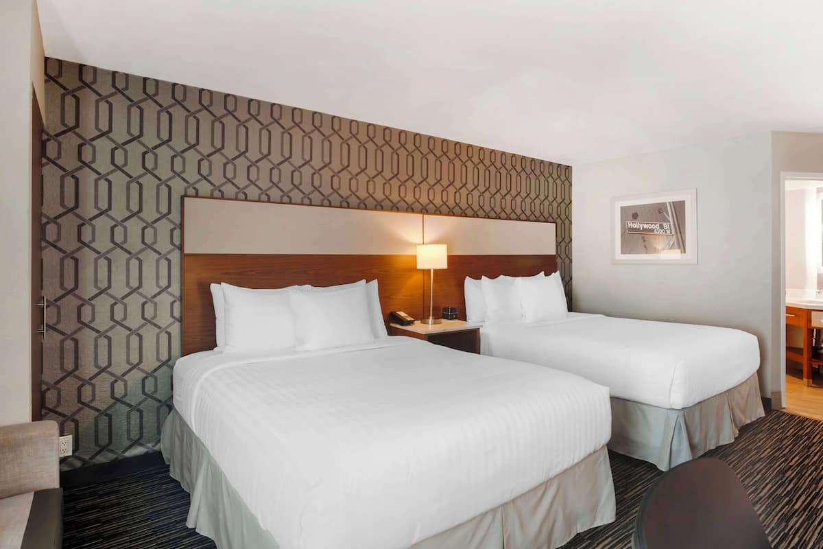 A hotel room with two beds, a patterned wall, a nightstand with a lamp, and a framed picture on the wall.