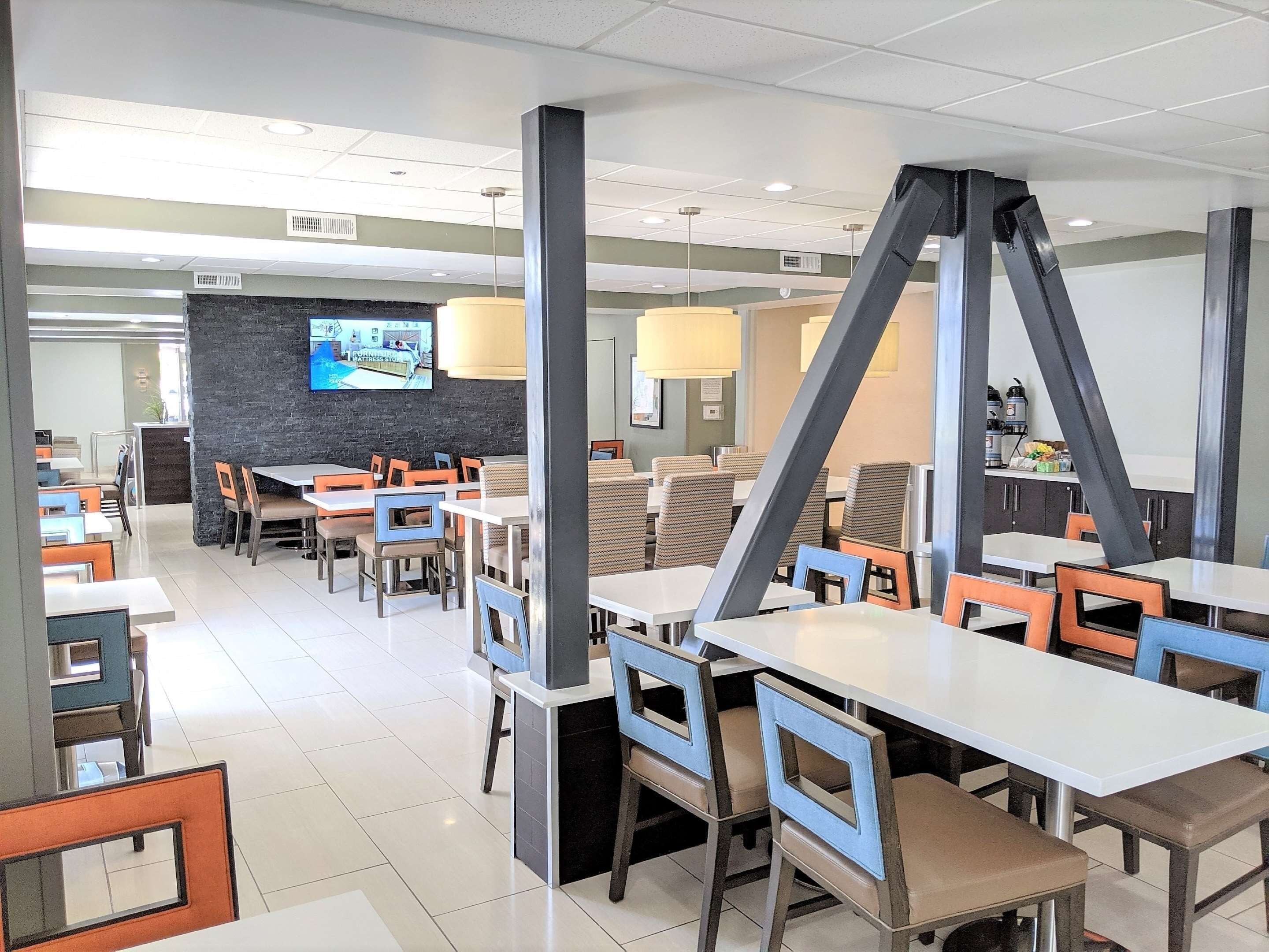 Un restaurante moderno con mesas y sillas blancas, una pared de acento negra y una pantalla de televisión mostrando una tienda de muebles.