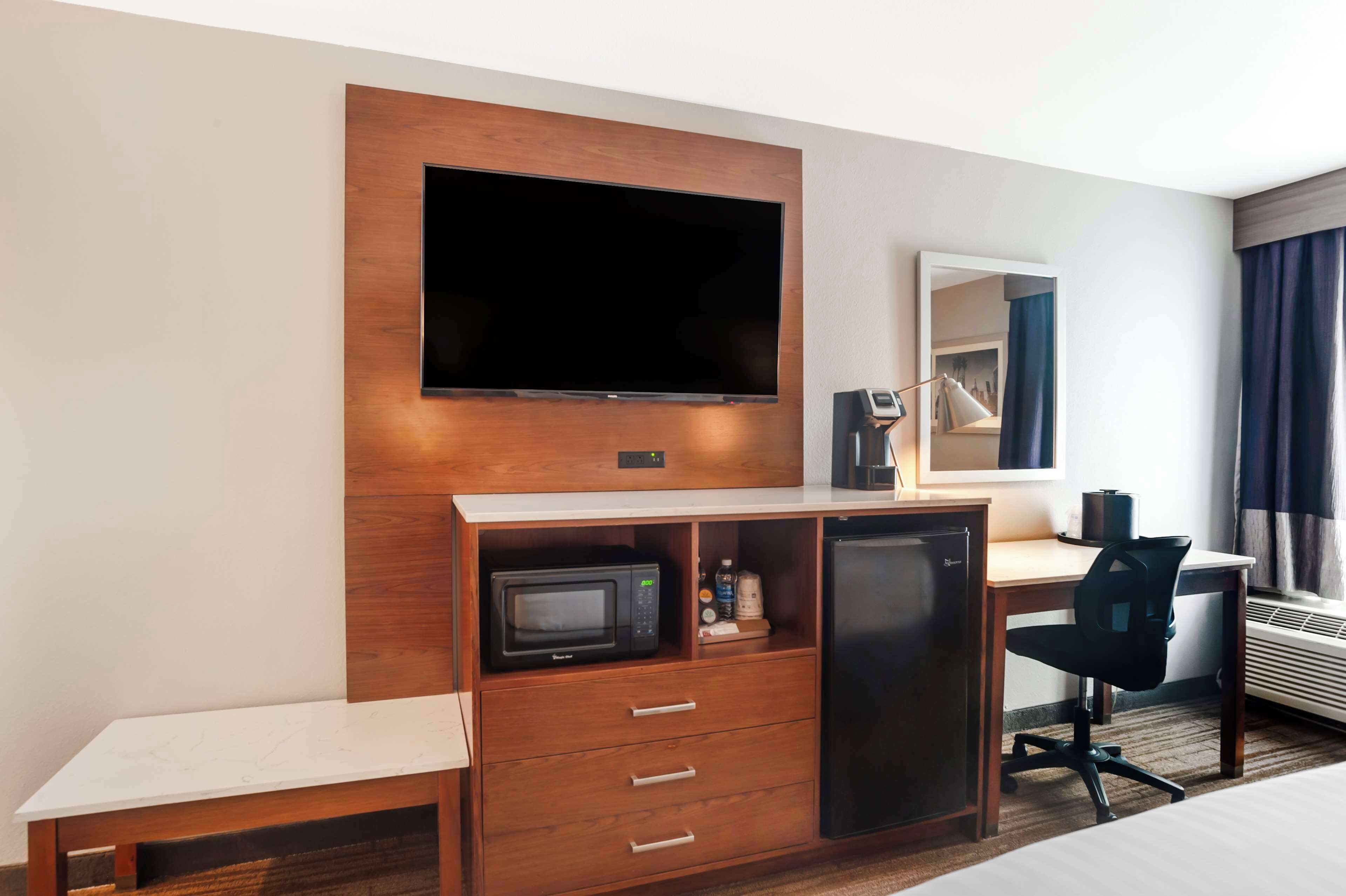 Habitación de hotel con televisión de pantalla plana montada en un panel de madera, un escritorio con una silla, microondas, refrigerador pequeño y una mesita de noche.