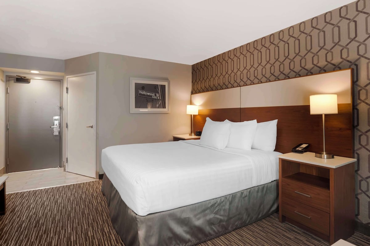 A hotel room with a large bed, two bedside tables with lamps, a wooden headboard, a framed picture on the wall, and a door.
