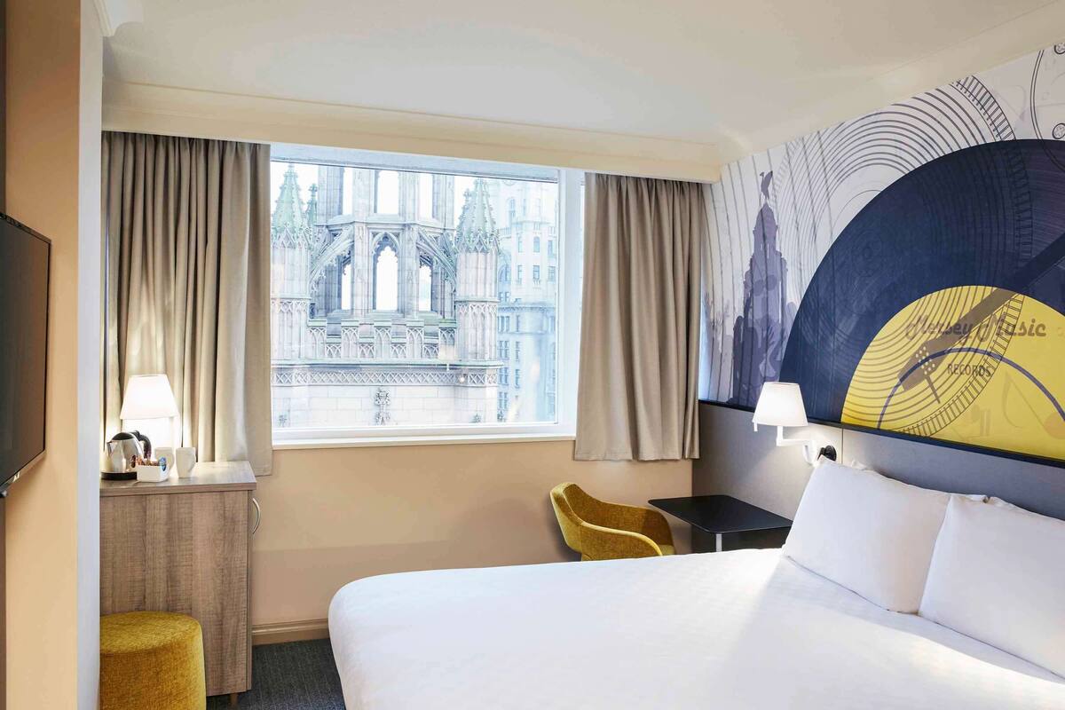 A hotel room with a large bed, a desk with a lamp, a chair, a small stool, and a view of a building through the window.