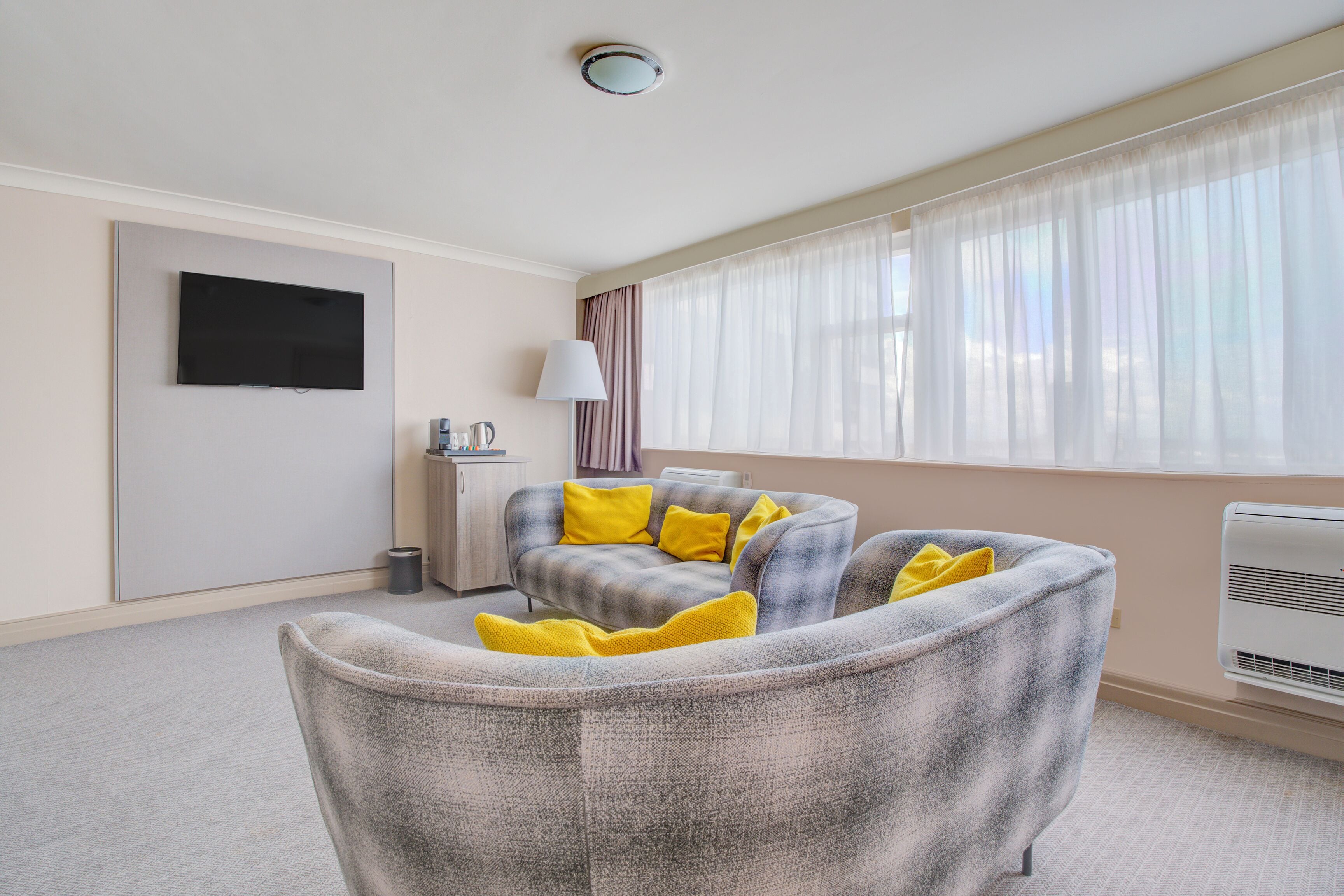 A modern living room with a grey sofa, yellow cushions, a flat-screen TV, a lamp, and a large window with sheer curtains.