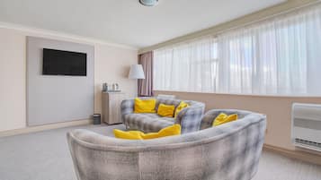 A modern living room with a grey sofa, yellow cushions, a flat-screen TV, a lamp, and a large window with sheer curtains.