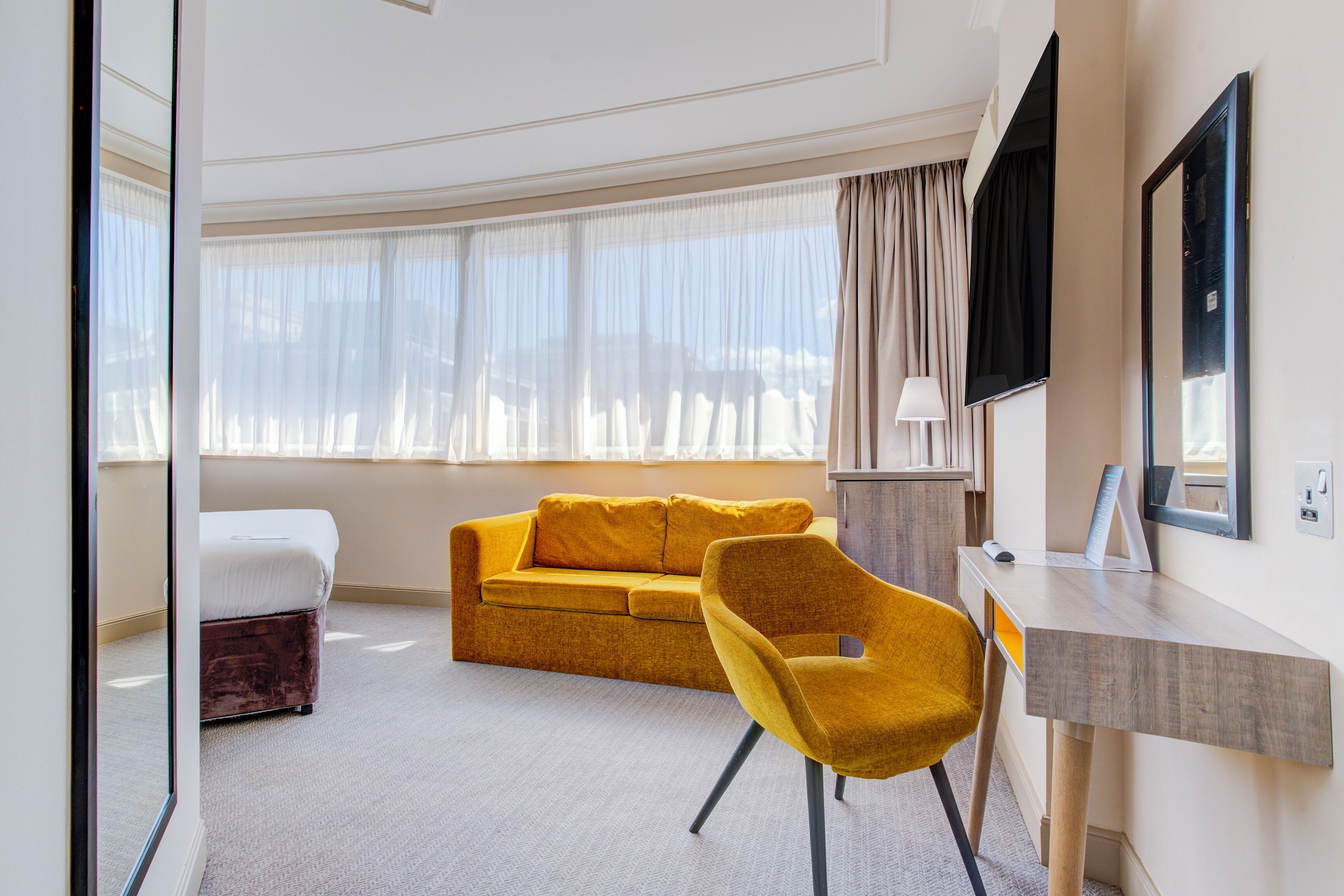 A hotel room with a yellow sofa, a yellow armchair, a desk, a TV, and a window with curtains.