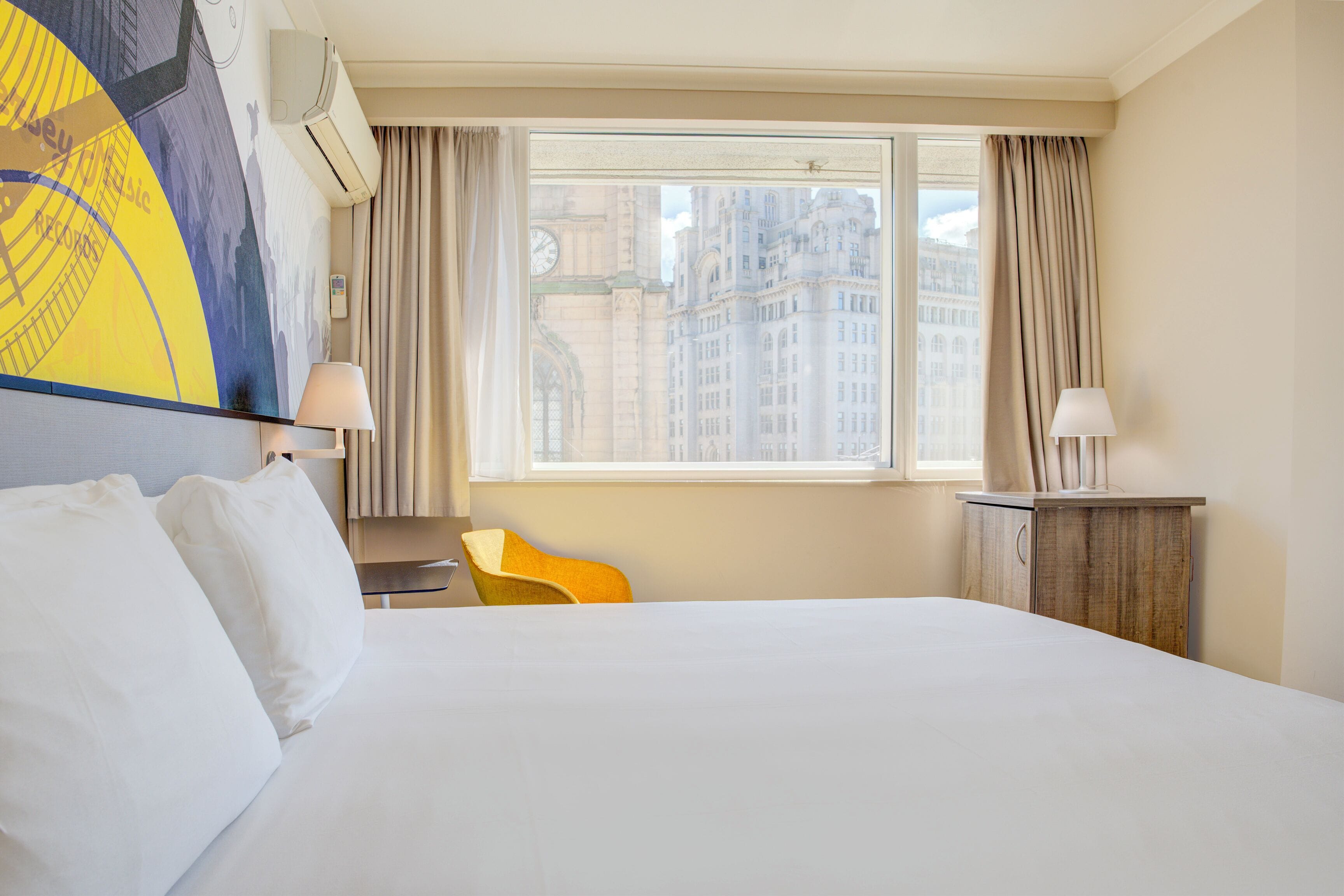 A hotel room with a large bed, a desk, a chair, and a view of city buildings through the window.