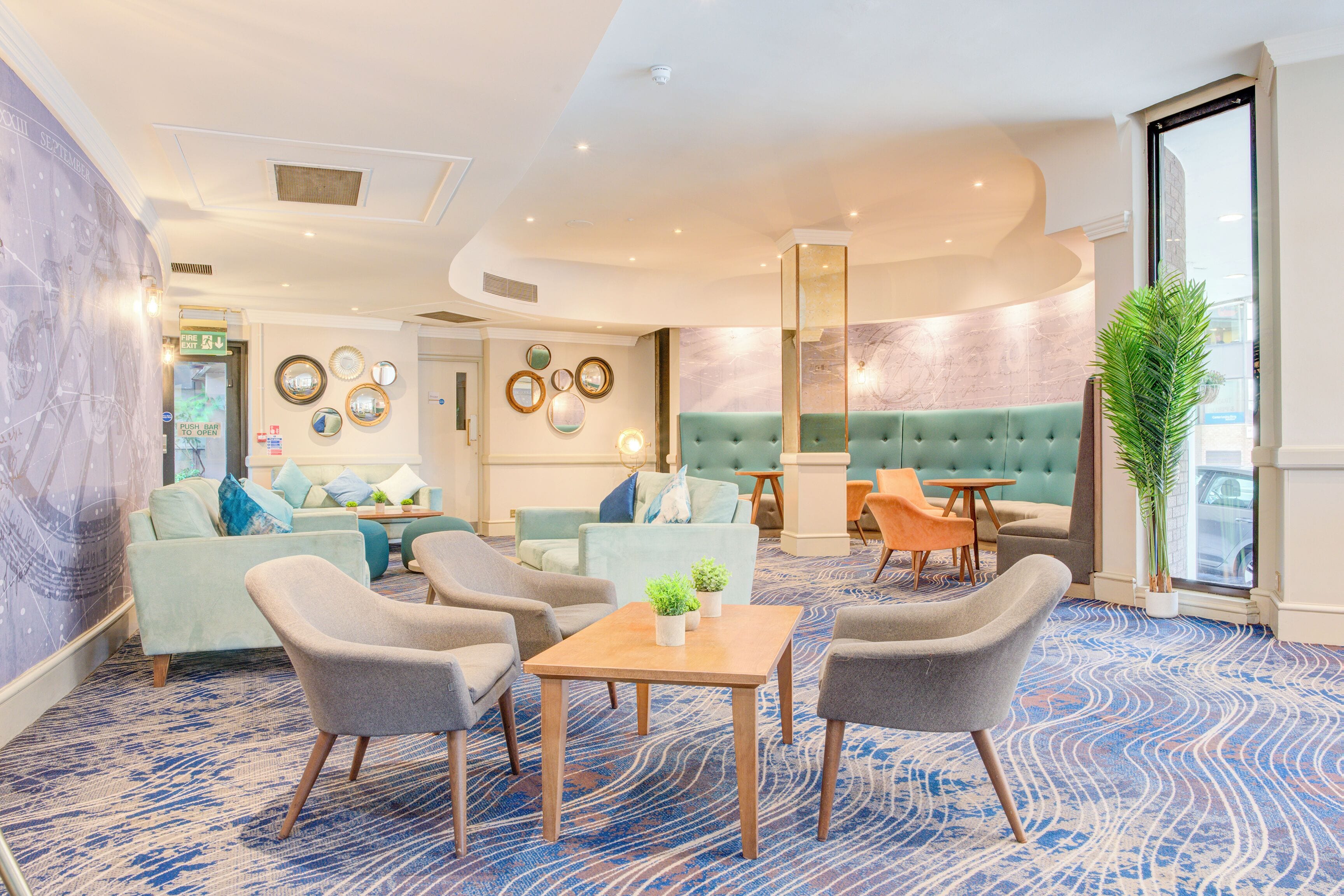 Un hall d’hôtel avec un tapis à motifs bleus, un canapé vert rembourré et une table basse en bois sur laquelle sont posées deux plantes en pot.