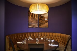 A dining area with a round table and tufted bench, illuminated by a pendant light fixture, surrounded by framed photographs and dark purple walls.