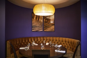 A dining area with a round table and tufted bench, illuminated by a pendant light fixture, surrounded by framed photographs and dark purple walls.