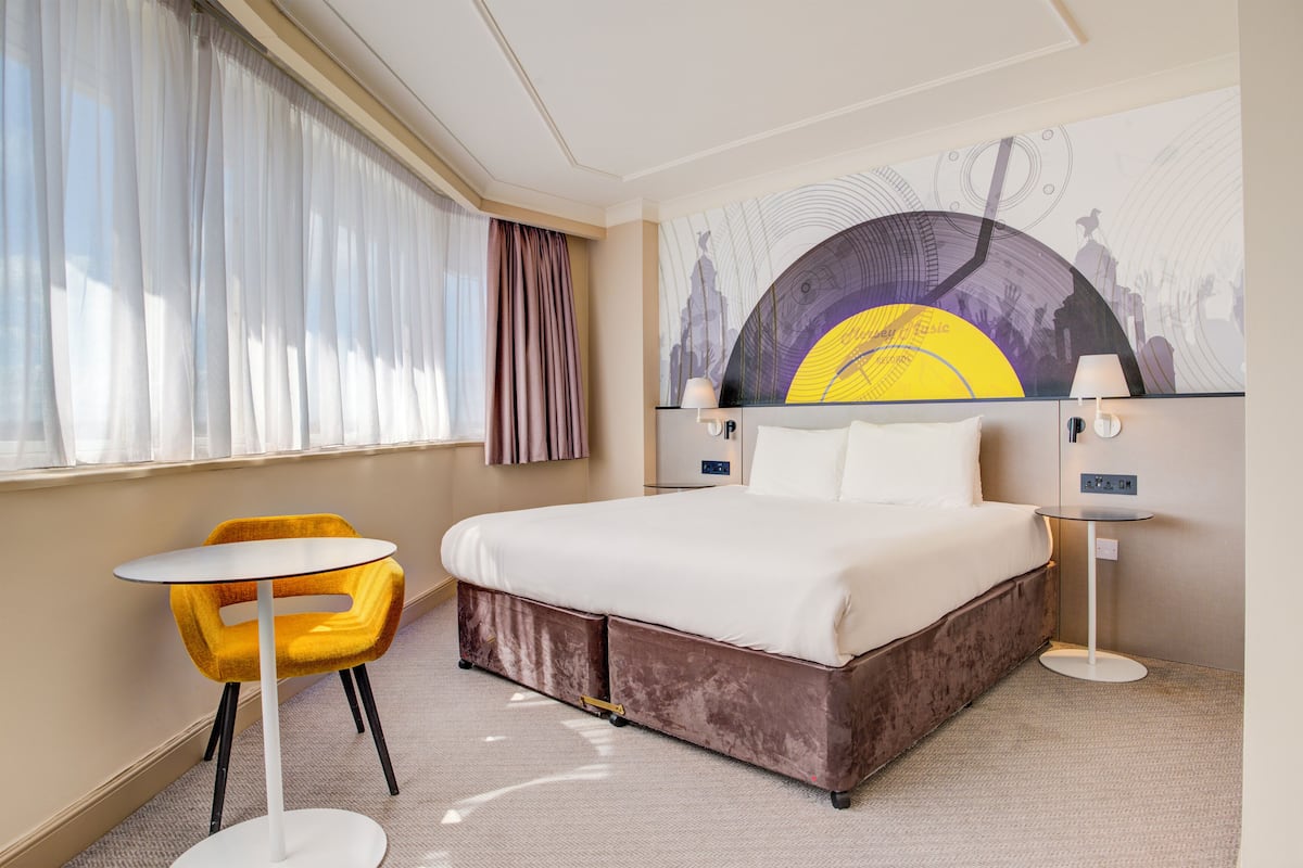A hotel room with a bed, a small table, a chair, and a large wall mural.