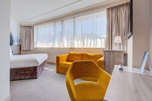 Habitación de hotel con una cama, un sillón amarillo, un sofá, un escritorio con lámpara y un ventanal con cortinas.