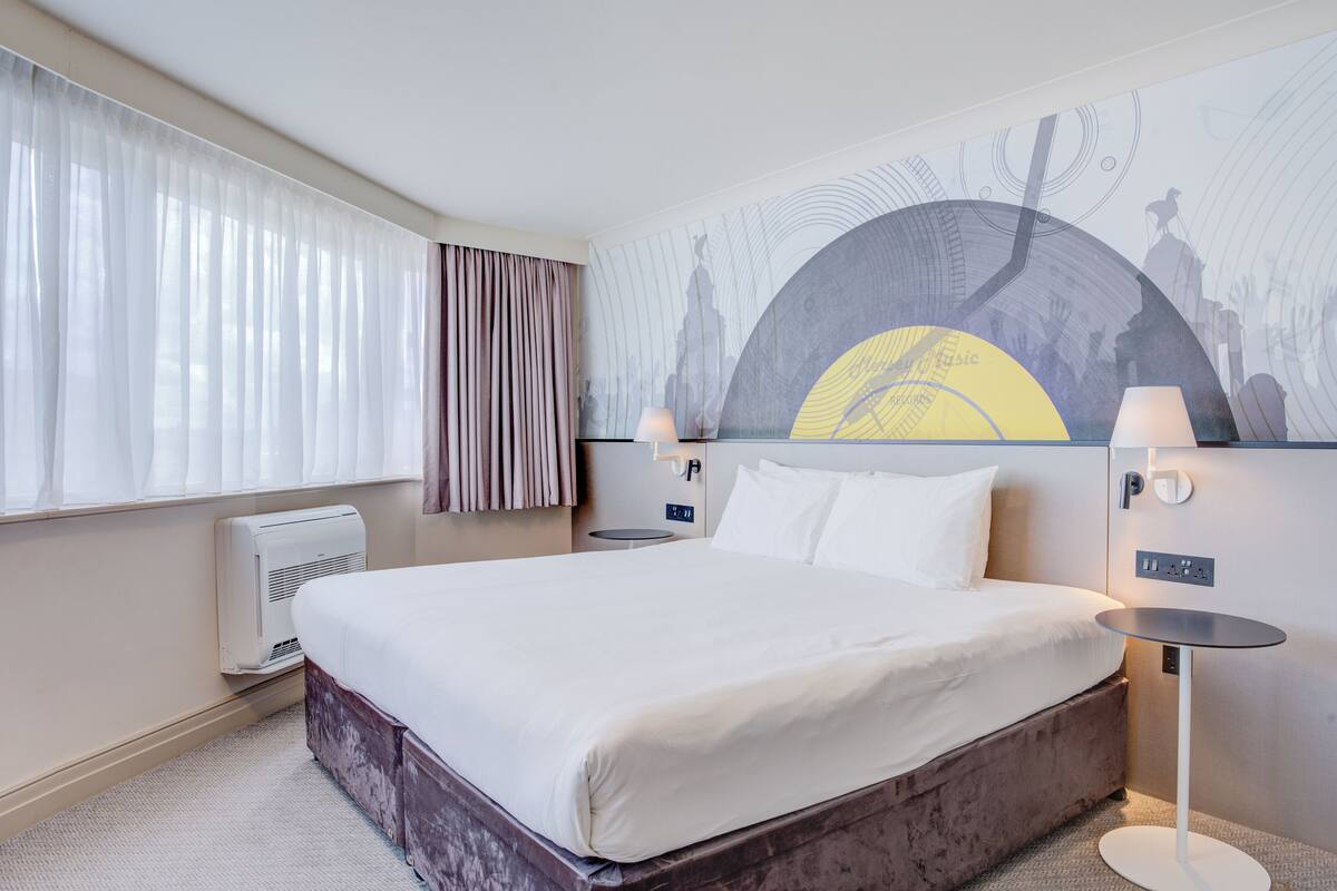 A hotel room with a large bed, two bedside lamps, a wall-mounted air conditioner, and a large abstract painting.