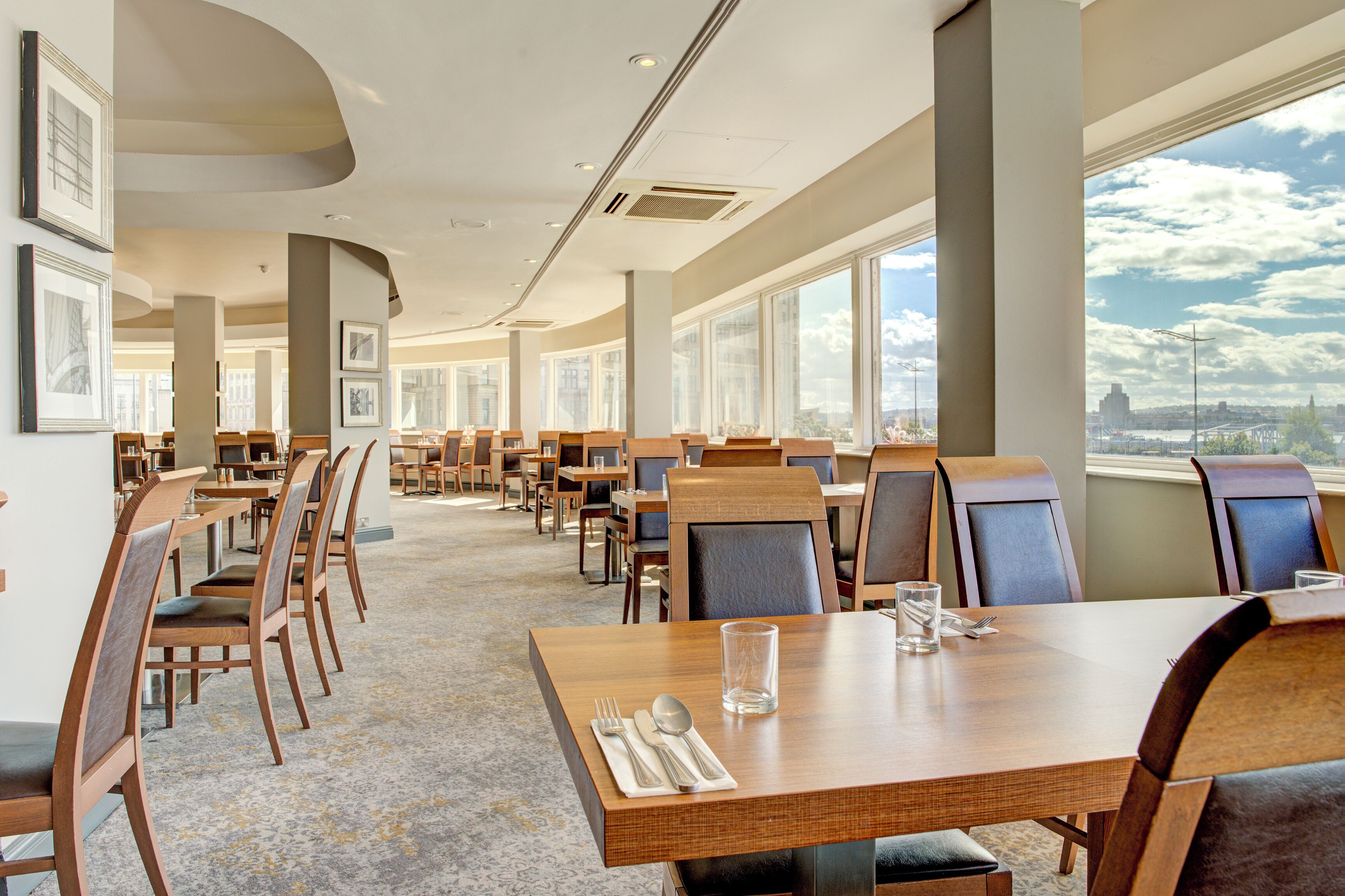 A spacious dining area with multiple tables and chairs, large windows offering a view of the cityscape, and framed artwork on the walls.