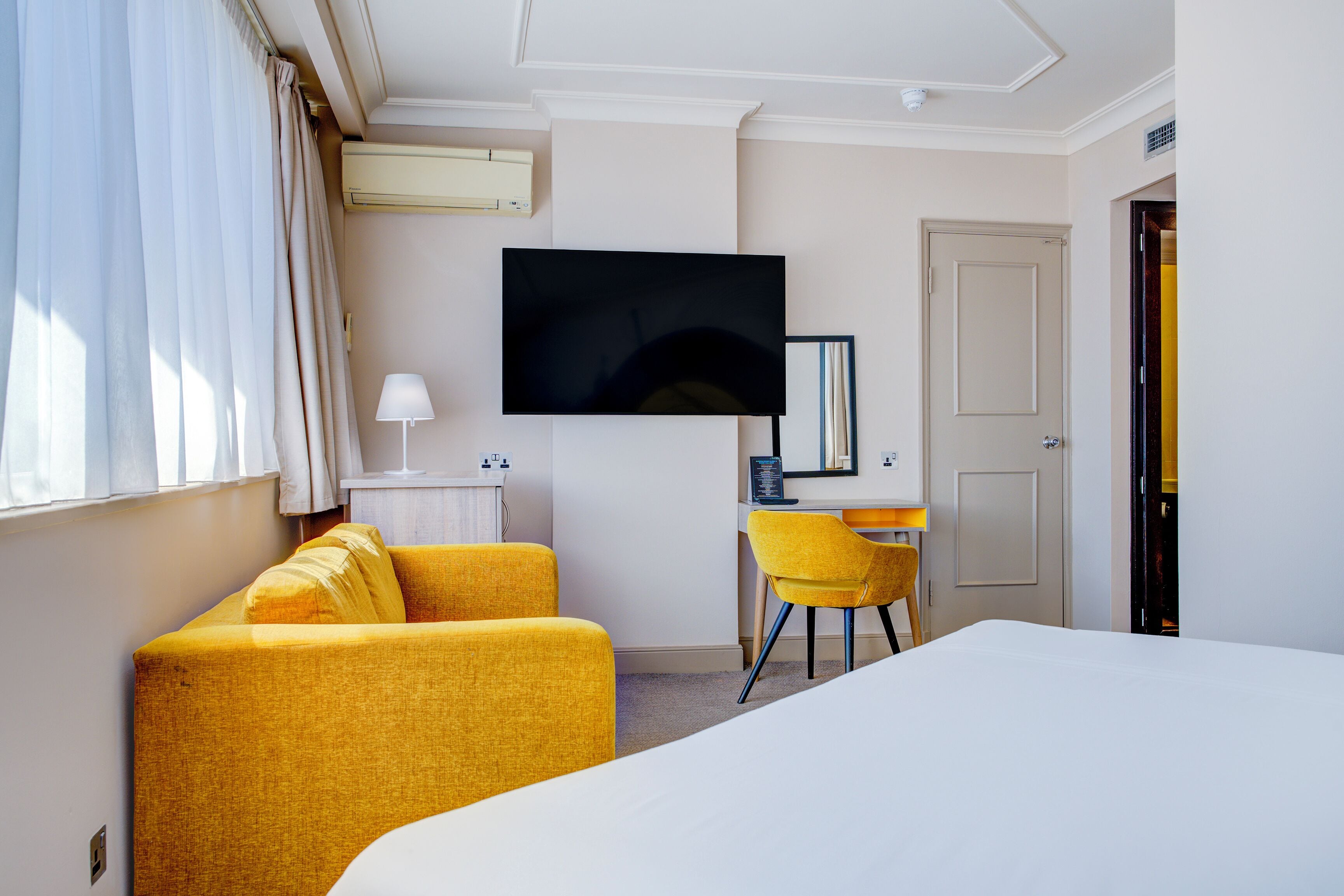 A hotel room with a bed, a desk with a chair, a TV mounted on the wall, and a door.
