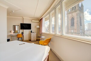 Habitación de hotel con cama, escritorio, silla, televisor y ventana con vista a un edificio histórico.