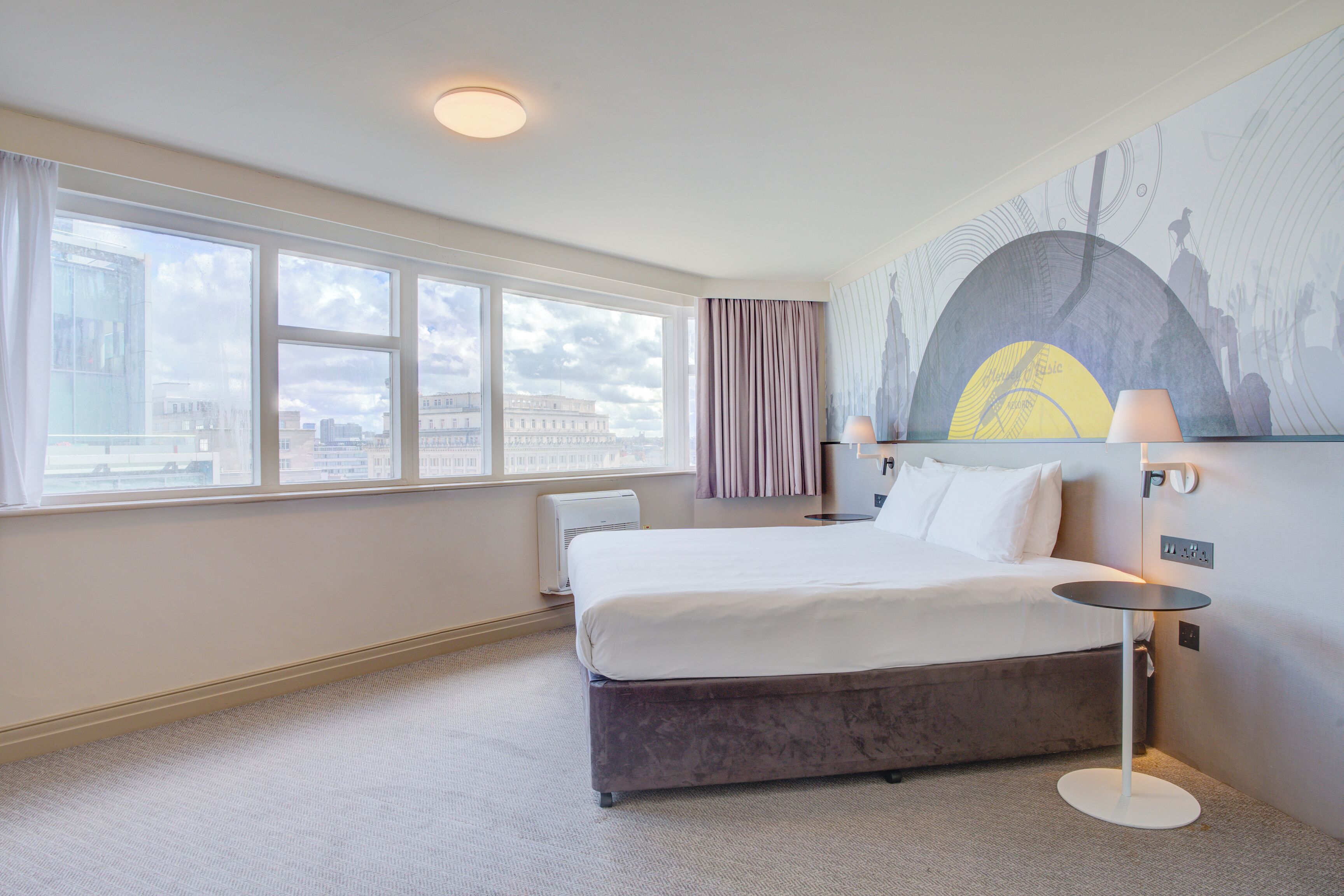 A hotel room with a large bed, a bedside table, a lamp, and a cityscape mural on the wall.