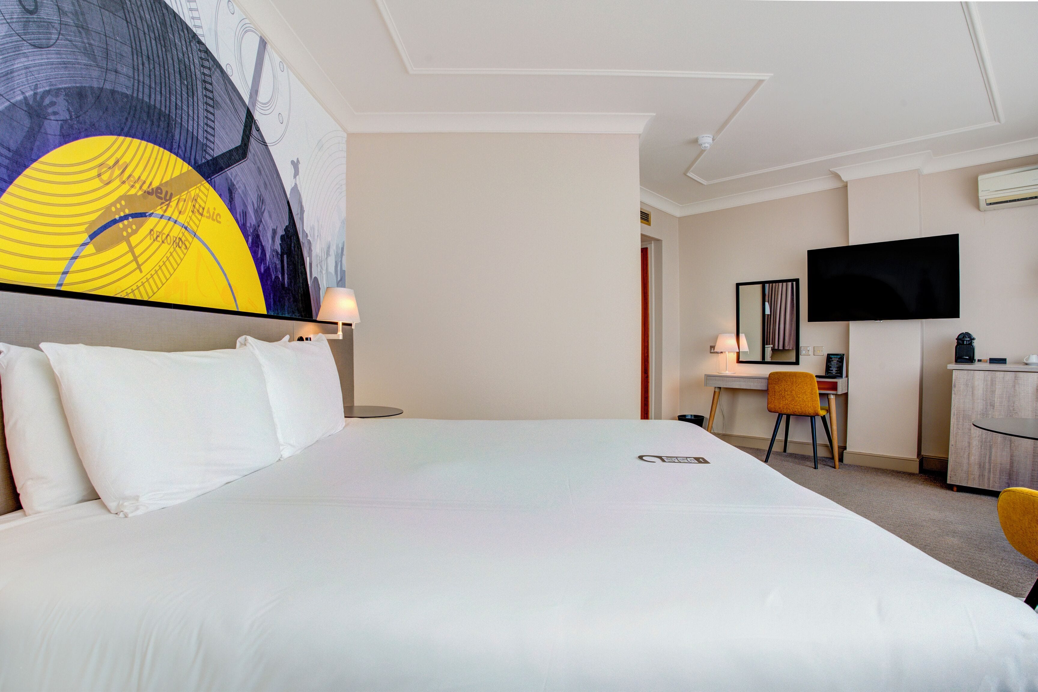 A hotel room with a large bed, a desk with a chair, a television, and a wall-mounted air conditioner.