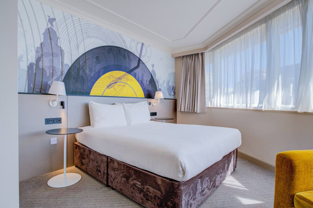 A hotel room with a large bed, two bedside tables, a wall mural, and a window with curtains.