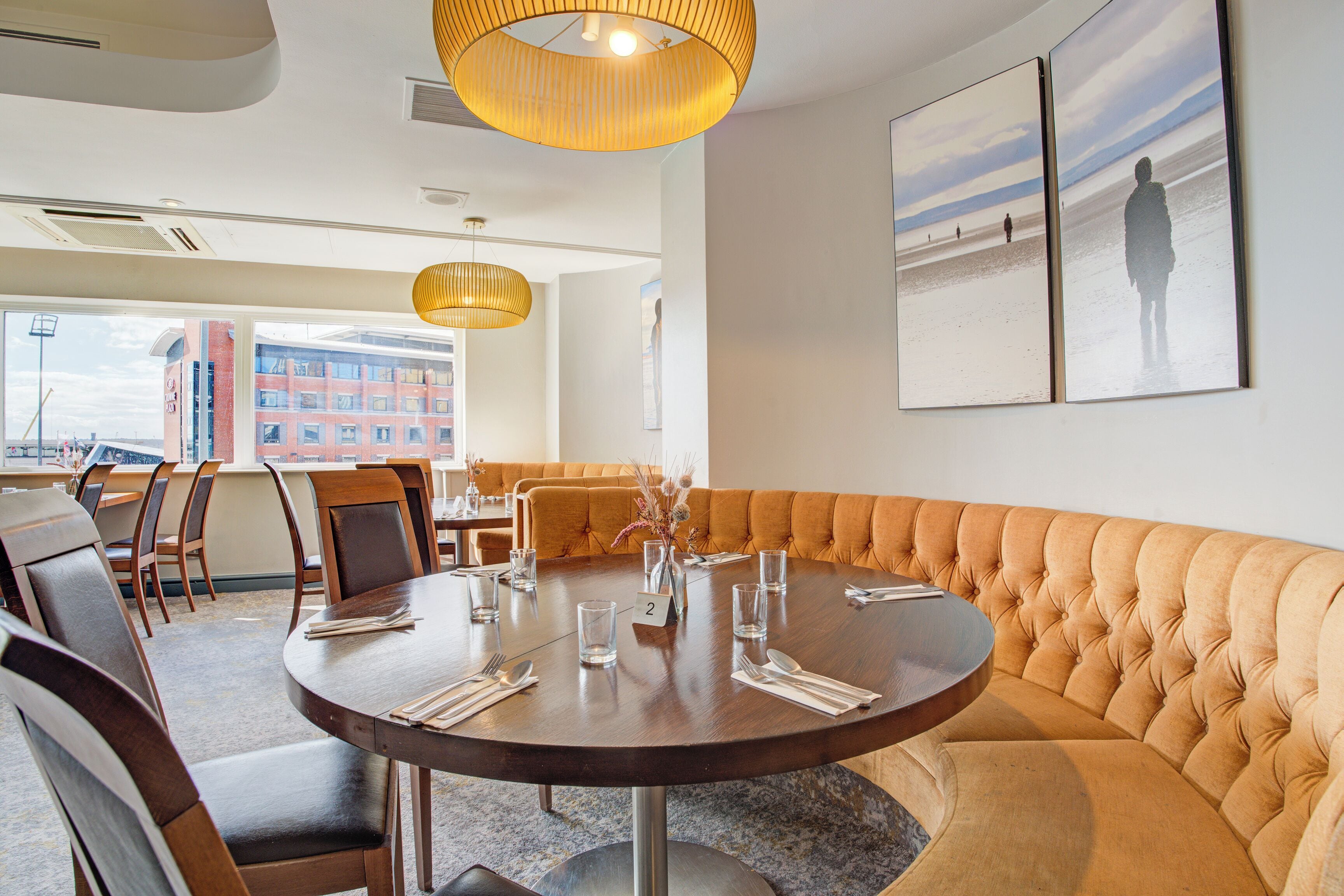 A dining area with a round table, chairs, and a cushioned bench. There are two framed pictures on the wall, a vase with flowers, and a number '2' on the table.