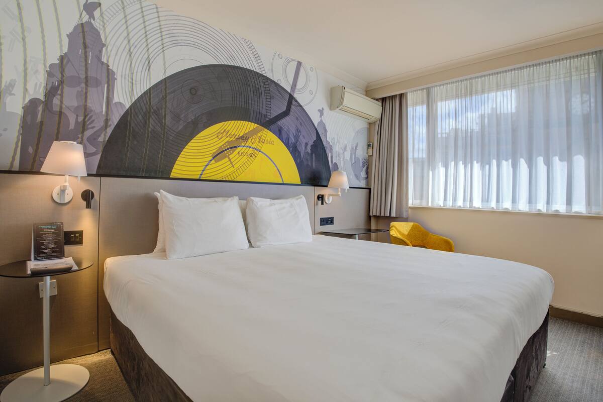 A hotel room with a large bed, a wall-mounted air conditioner, a small table with a lamp, and a yellow chair.