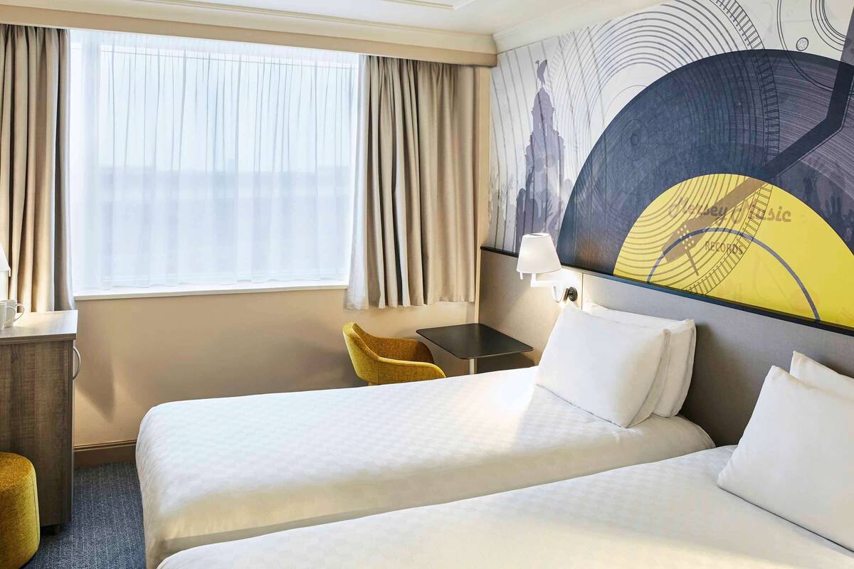 A hotel room with two beds, a desk, a chair, a lamp, and a large wall mural.
