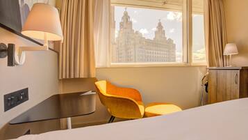A hotel room with a bed, a desk, a chair, a lamp, a window with a view of a building, and a wardrobe.