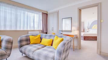 A hotel room with a grey plaid sofa, a desk with a lamp, a bed, and a large window with sheer curtains.