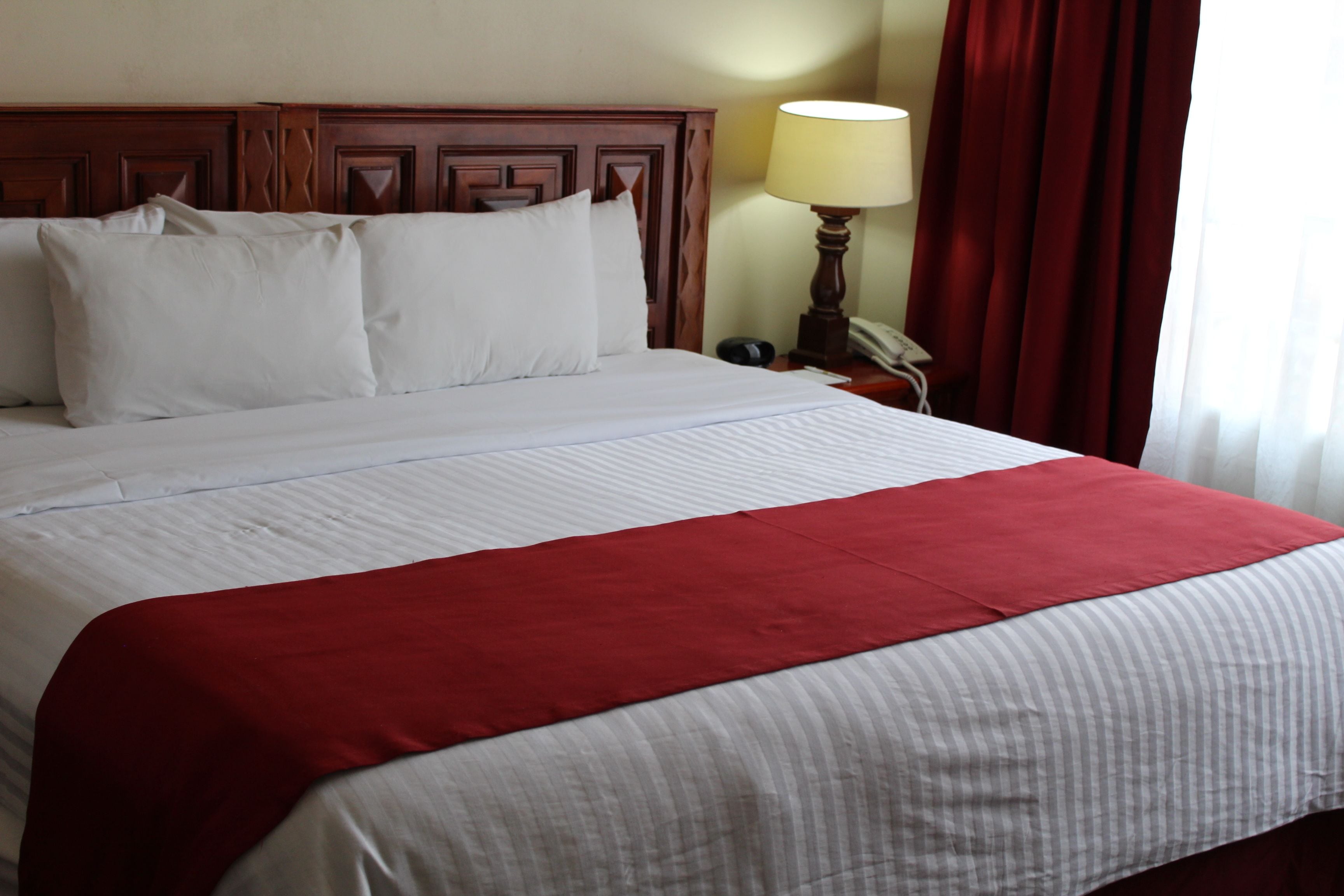 A neatly made bed with white and red bedding, a wooden headboard, a bedside lamp, and a red curtain.