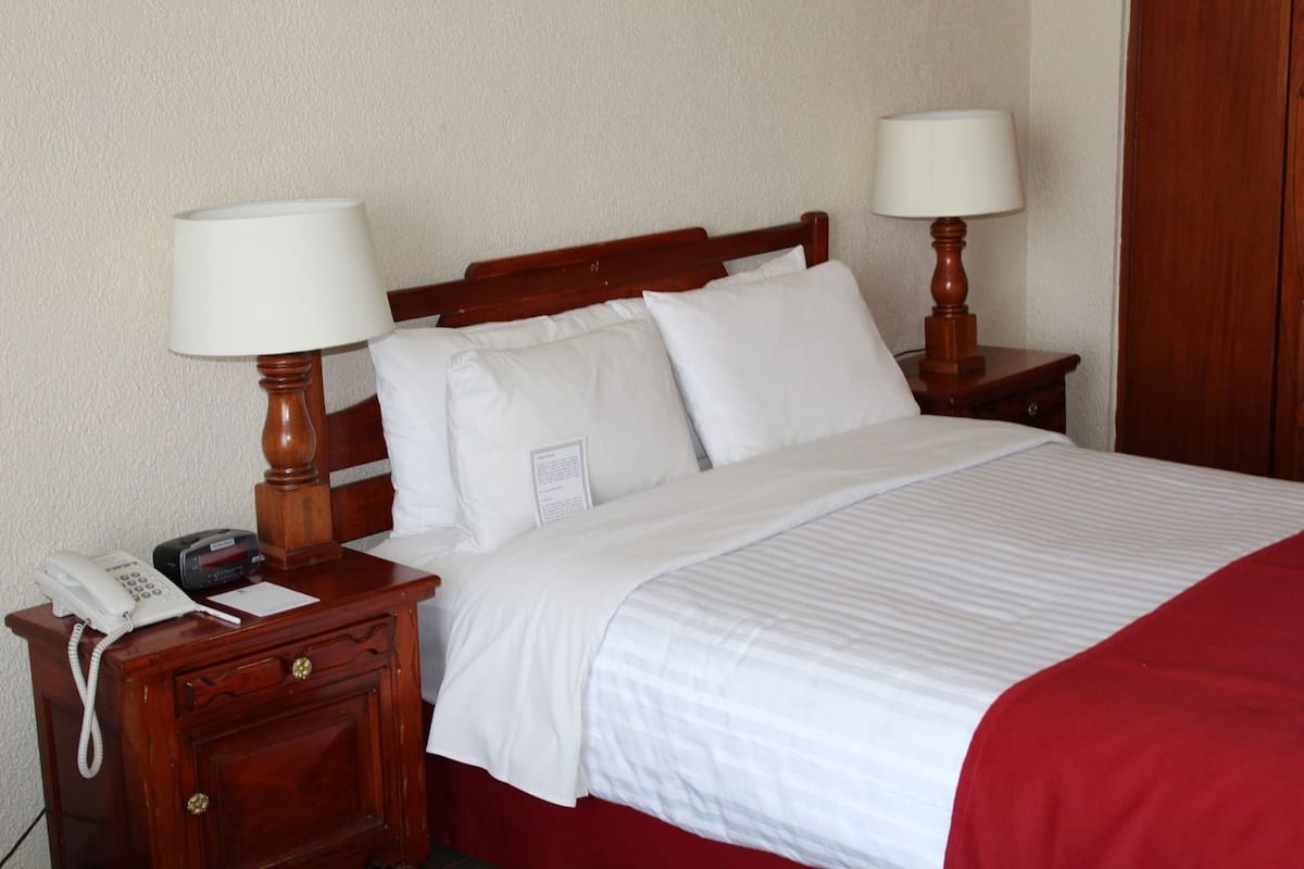 Una cama bien hecha con sábanas blancas y una manta roja, dos lámparas de noche, una mesita de noche de madera con un teléfono, y una lámpara de pared.