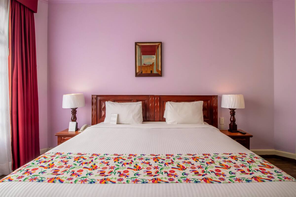 A hotel room with a large bed, two bedside tables with lamps, a wooden headboard, and a painting on the wall.