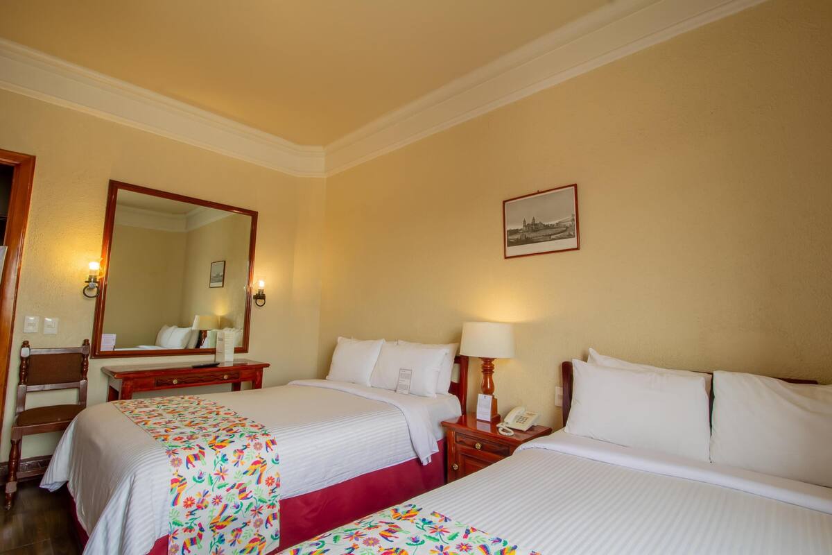 A hotel room with two beds, a wooden nightstand with a lamp, a desk with a chair, a large mirror, and a framed picture on the wall.