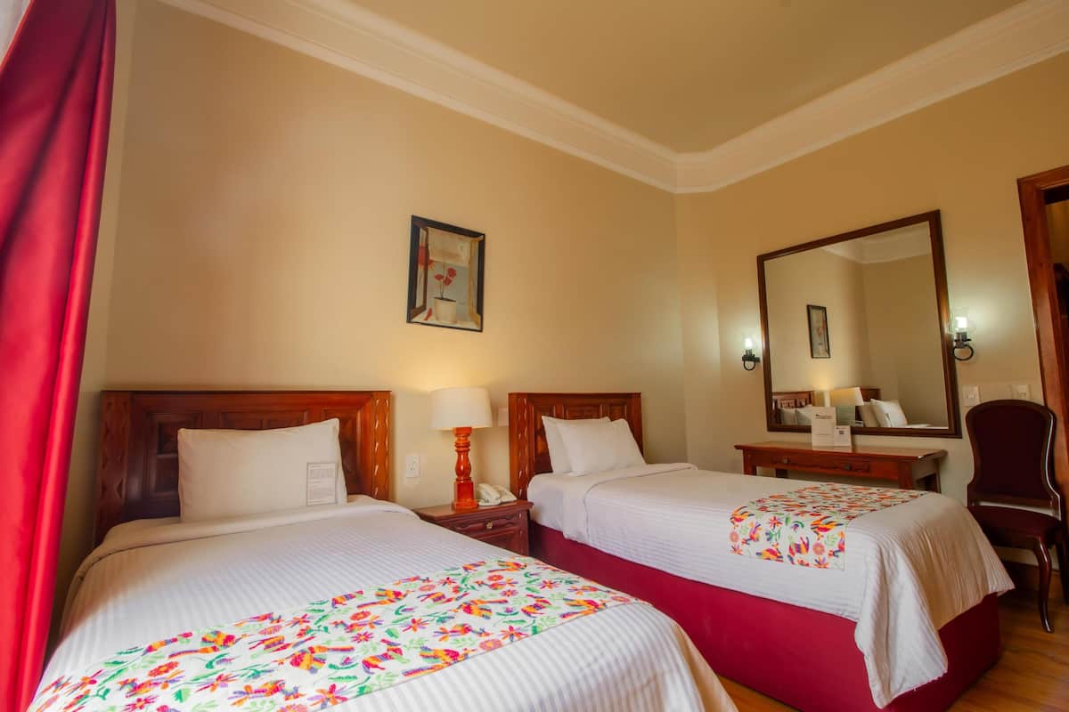A hotel room with two beds, a wooden headboard, a nightstand with a lamp, a mirror, and a chair.