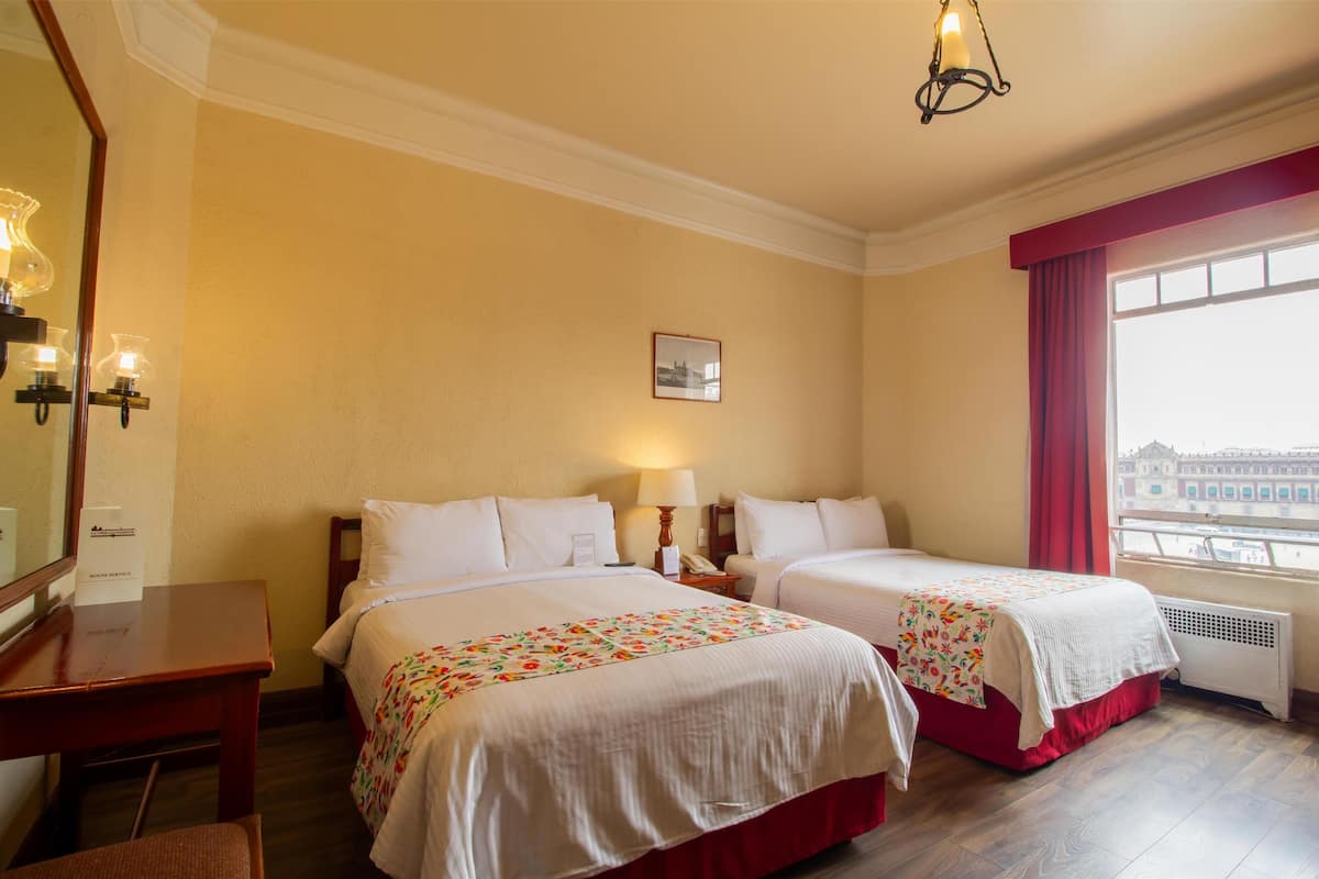 A hotel room with two beds, a desk, a chair, a window with a view, and a ceiling light fixture.