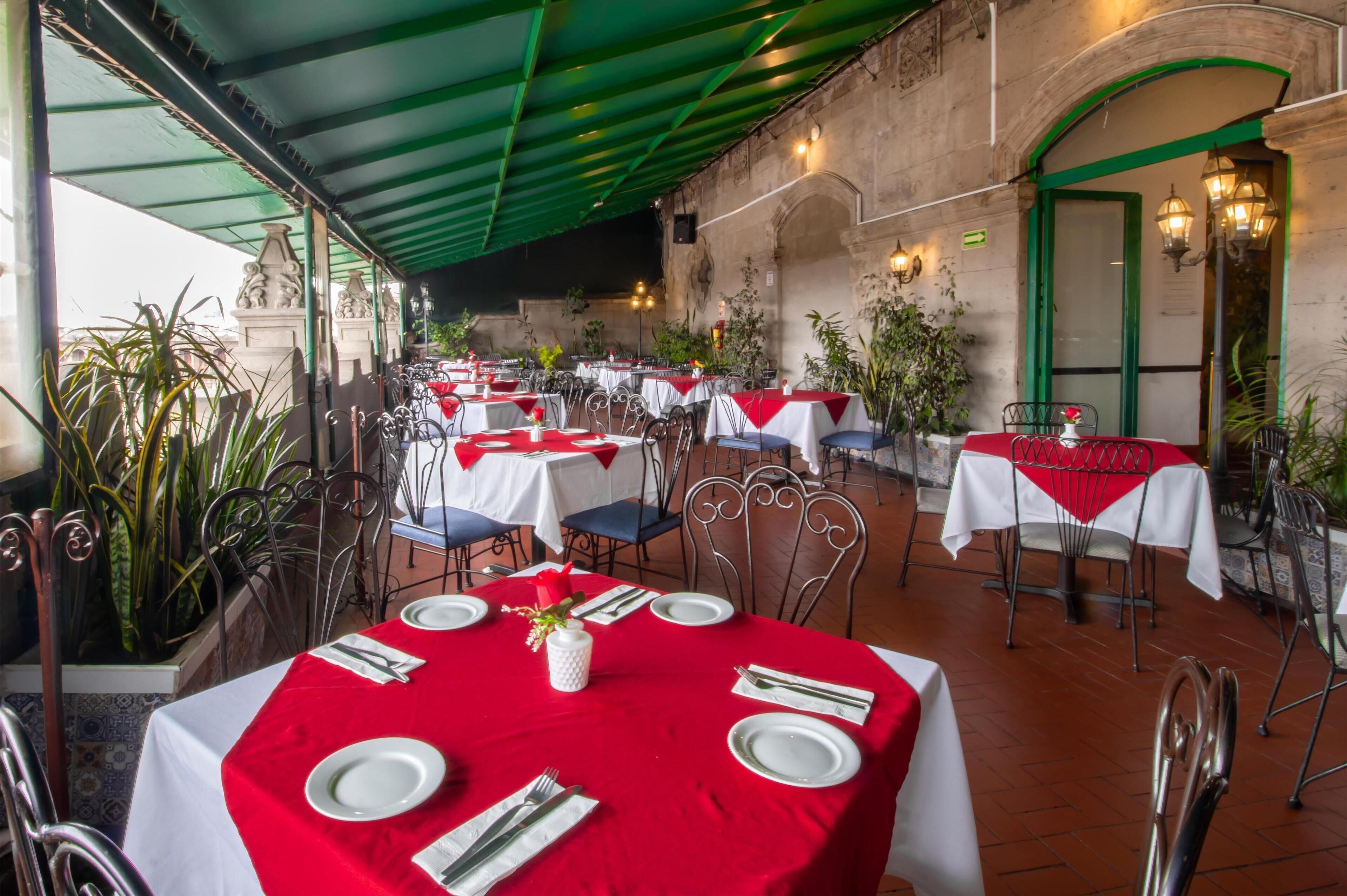An outdoor dining area with tables set for two, featuring red tablecloths and white plates.
