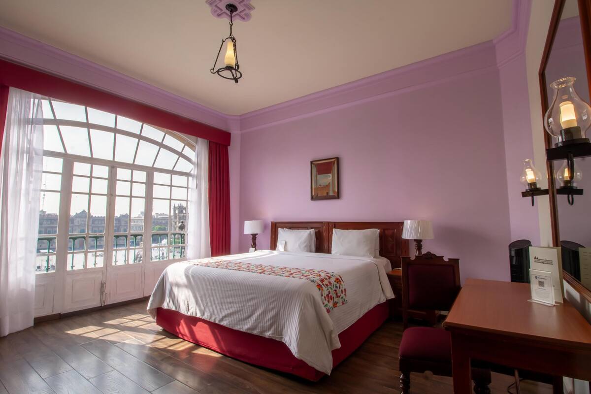 A hotel room with a large bed, a desk, and a view of a cityscape through large windows.