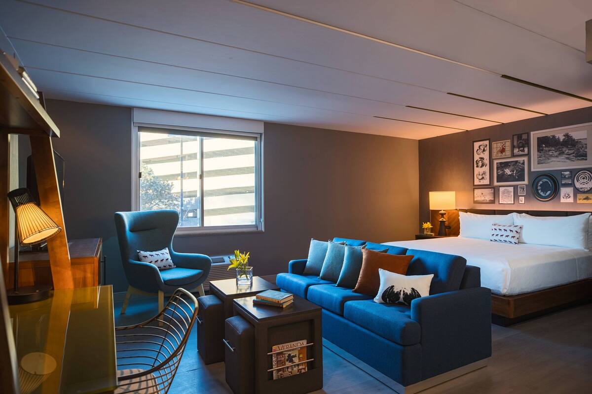 A modern hotel room with a large bed, a blue sofa, a desk with a lamp, and a window providing natural light.
