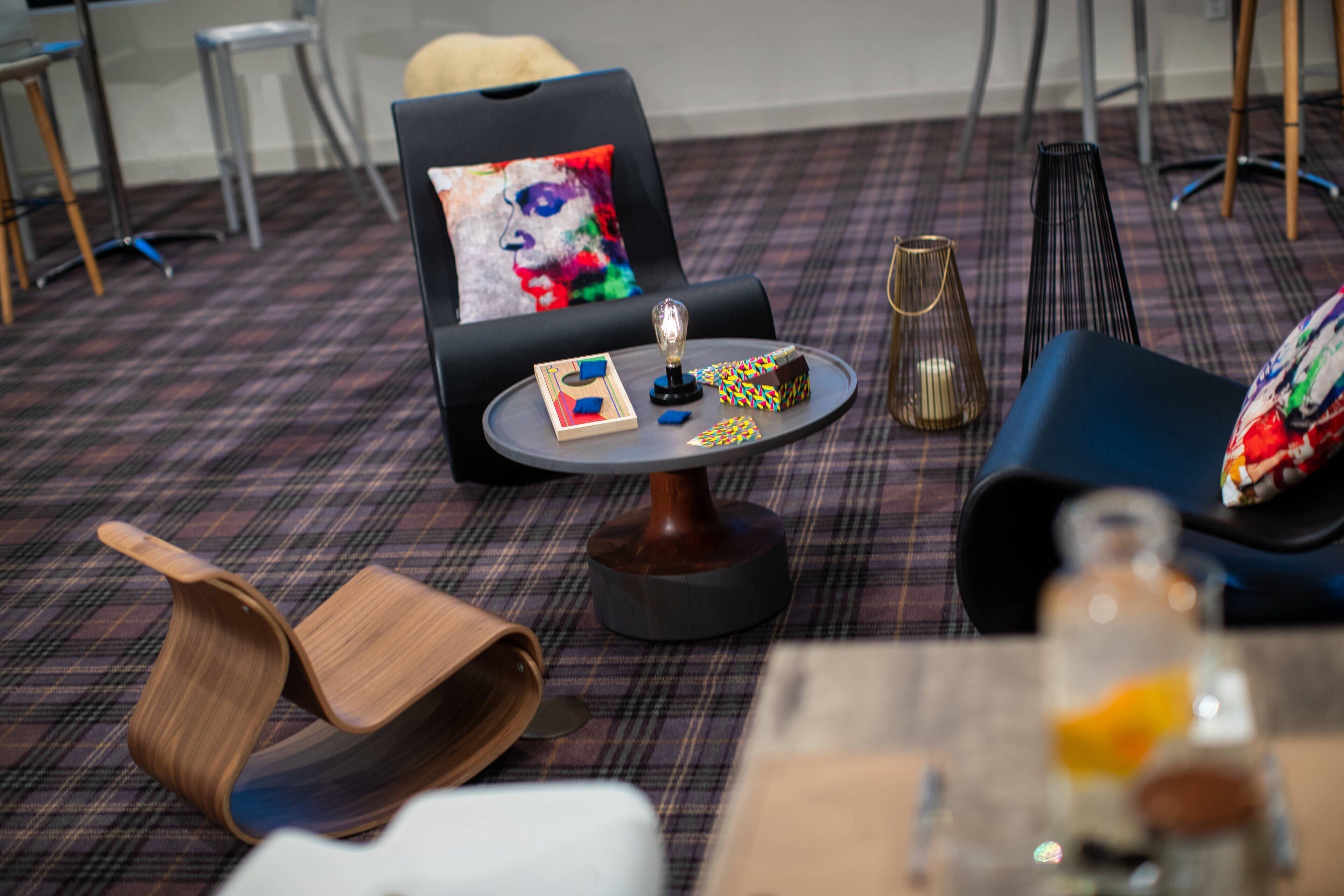 Un área de descanso moderna con alfombra escocesa, un sillón negro con un cojín de colores, una pequeña mesa redonda con una lámpara y una botella de vidrio sobre una mesa de madera.