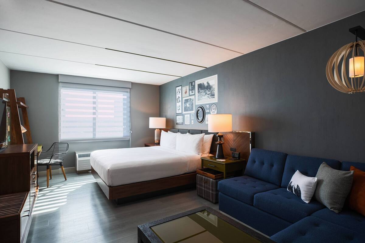 A modern hotel room with a large bed, a blue sofa, a desk, and a wall with framed pictures.
